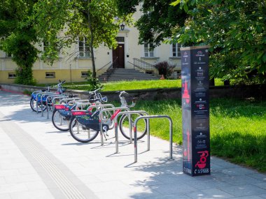 Şehir merkezinde modern binanın yanında renkli bisikletler ve yeşil peyzaj düzenlenmiş şehir bisikleti otoparkı.