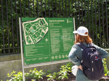 Beyaz şapkalı kadın turist, yeşil çitin yanında mor sırt çantası taşırken parkın bilgilerini inceliyor.