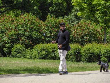 Yeşilliklerle çevrili yeşil park yolunda tasmalı küçük siyah köpeği gezdiren kişi.