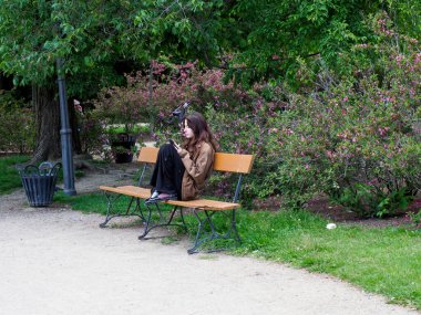 Parktaki bankta kitap okuyan kadın yemyeşil bitki örtüsüyle çevrili. Mükemmel bir açık hava rahatlama anı yaratıyor.