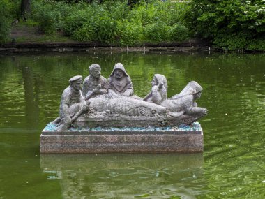 Üç figürlü taş heykel yemyeşil parktaki sakin gölette huzur içinde oturuyor.