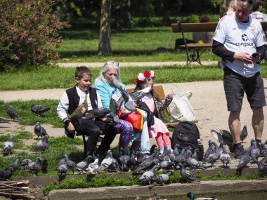 Torunları olan yaşlı bir çift şehir parkında güvercinleri beslerken neşeli aile anları yaratıyor.