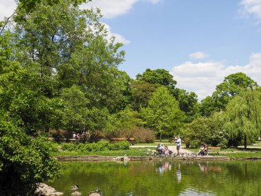 Olgun ağaçlarla dolu yemyeşil park manzarası ve dingin göl kenarında dinlenen insanlar.