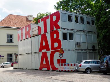Ön planda cephe ve inşaat malzemeleri üzerine büyük kırmızı harflerle modern sanayi binası
