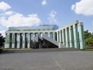 İş bölgesinde geometrik mimarisi ve beton plazası olan modern yeşil cam ofis binası