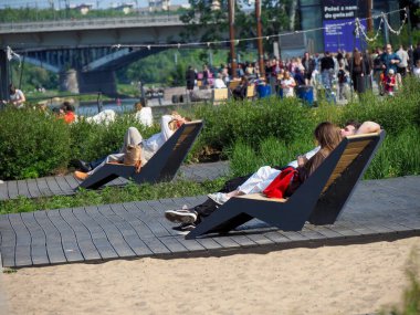 İnsanlar Varşova Vistula nehir kıyısı yakınlarındaki ahşap şezlonglarda dinlenirken köprü arkasında kalabalık toplanıyor.