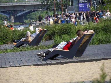 Turistler, Varşova 'daki Vistula nehrinin kıyısındaki ahşap şezlonglarda güneşli bir öğleden sonranın keyfini çıkarıyorlar.