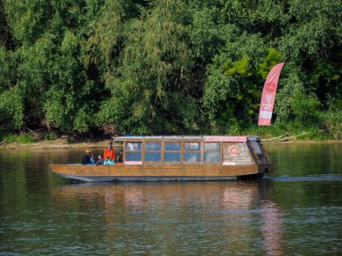 Geleneksel ahşap yolcu botu nehir kenarındaki iskeleye yanaştı. Pembe bayrak ve orman zeminiyle.