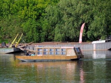 Tahta yolcu feribotu limana demirlemiş sarı direkler ve yeşil orman manzaralı.