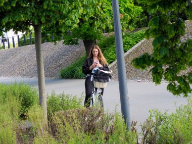 Modern şehir parkında duran bisikletli genç kadın yemyeşil bitki örtüsüyle çevrili.
