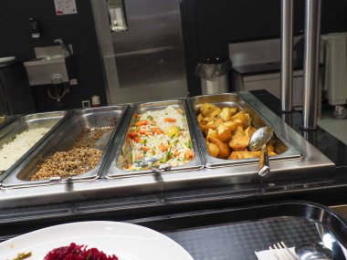 Profesyonel kafeterya büfesi. Birden fazla yiyecek kabı var. Çeşitli sıcak yemek çeşitlerini gösteriyor.