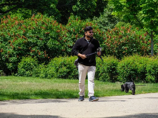 Orta yaşlı bir adam, yemyeşil bitki örtüsüyle çevrili güneşli park yolunda tasmalı küçük siyah köpeği gezdiriyor.