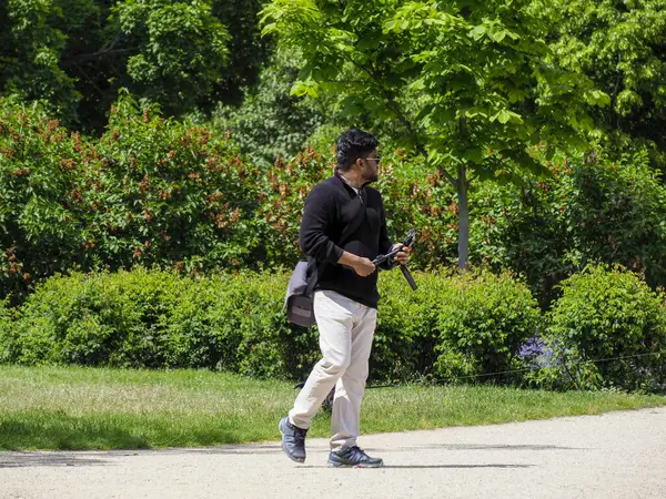 Koyu renk elbiseli olgun bir adam gündüz vakti yeşil park ortamında küçük siyah bir köpekle yürüyüp gidiyor.