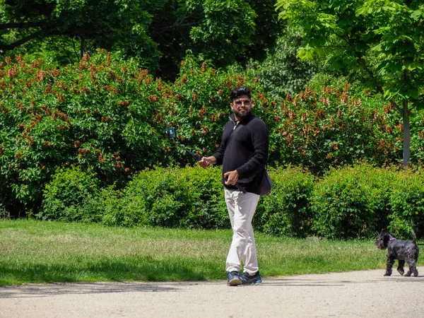 Yeşilliklerle çevrili yeşil park yolunda tasmalı küçük siyah köpeği gezdiren kişi.