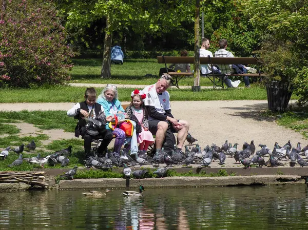 Çocuklu aileler, sabah güneşinin tadını çıkarıyorlar. Yansıtıcı su birikintisi kenarında güvercin besliyorlar.