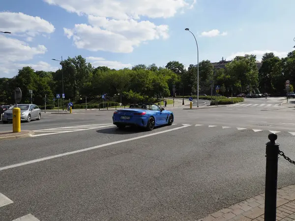 Geniş, boş bir otopark, mavi spor arabası ve bulutlu gökyüzünün altında yeşil ağaçları var.