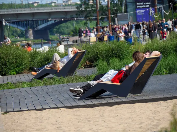 İnsanlar Varşova Vistula nehir kıyısı yakınlarındaki ahşap şezlonglarda dinlenirken köprü arkasında kalabalık toplanıyor.