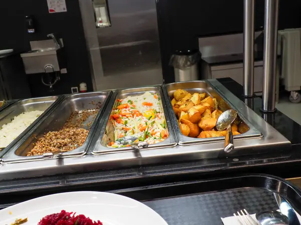 Profesyonel kafeterya büfesi. Birden fazla yiyecek kabı var. Çeşitli sıcak yemek çeşitlerini gösteriyor.