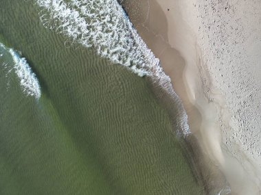Okyanus dalgalarının yukarıdan aşağıya doğru görüntüsü kumlu kıyıya ulaşırken karmaşık köpük desenleri oluşturuyor.