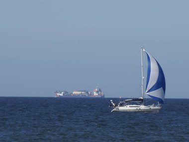 Ufukta sanayi gemileriyle Baltık Denizi sularında mavi ve beyaz yelkenli yelkenliyle zarif bir yelkenli.