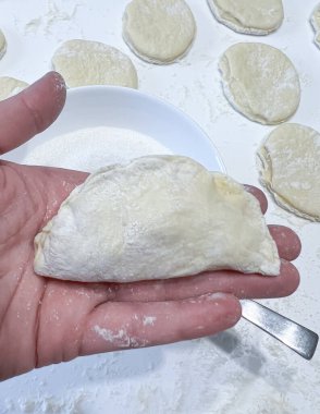 Kişi elinde masa hamurundan bir parça tutuyor ve geleneksel bir yemek pişirmeye hazırlanıyor. Mutfak gereçleri ve malzemeleri masada hazır.