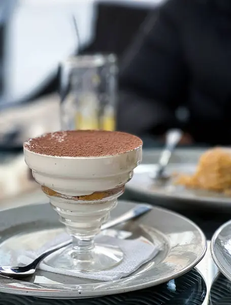 Masanın üzerinde bir tabak tiramisu servisi sunulur, kahve kaplı sünger keki ve kremalı mascarpone peyniri katmanları ile yapılan lezzetli tatlıyı gösterir.