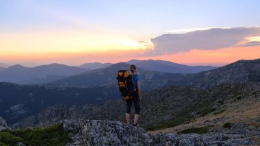Kadın yürüyüşçü, arkasında dağlar olan büyük bir kayanın üzerinde duruyor ve gün batımını izliyor..