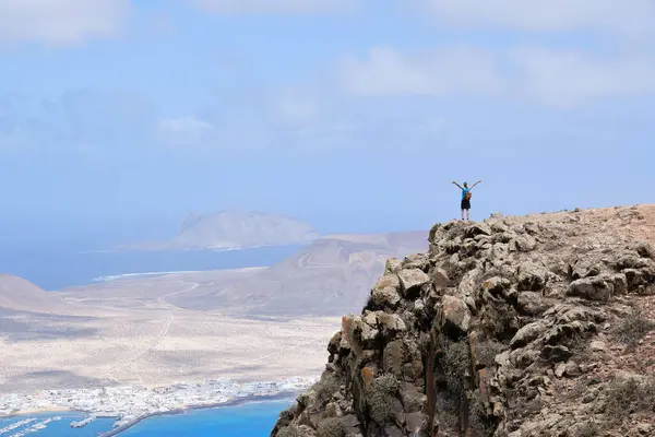 Kollarını bir tepenin üstüne kaldırmış bir kadın. Arka planda La Graciosa Adası var.