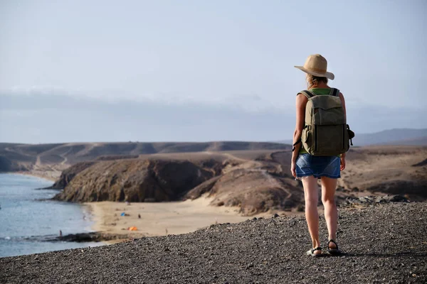 stock image Woman with hat and backpack, standing on top of a cliff and looking out to sea and beach at sunset. Travel and tourism.