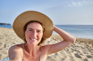 Hasır şapkalı neşeli bir kadın kumlu sahilde battaniyede dururken kameraya bakıyor ve mavi gökyüzüne karşı gün ışığında bulanık dalgalı denizin yanında selfie çekiyor.