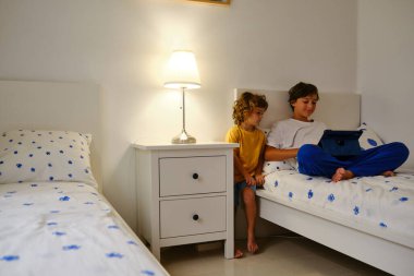 Şirin kıvırcık saçlı çocuk. Kardeşinin yanında duruyor. Yatağa uzanıyor ve yatmadan önce tablette film seyrediyor.