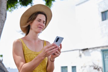 Yazlık kıyafetli sarışın kadın ve hasır şapkalı sokak kenarında duruyor ve güneş ışığında cep telefonuna bakıyor.