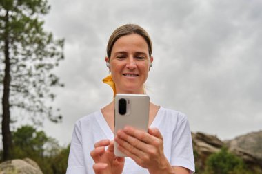 Taşrada bulutlu gökyüzüne karşı akıllı telefon kullanırken kulaklık takıp gülümseyen orta yaşlı kadın portresi