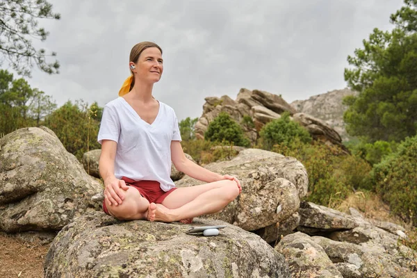 Full body of pensive female in casual clothes sitting on stone with easy pose yoga and closed eyes while meditating on rocky cliff with plants on cloudy day