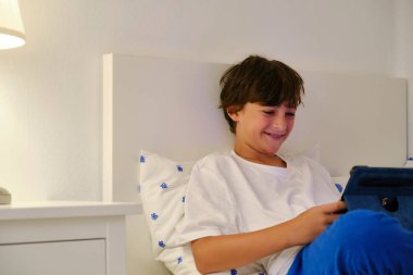 Beyaz tişörtlü ve kot pantolonlu mutlu genç çocuk tabletle yatağa uzanıp komik bir film izliyor.