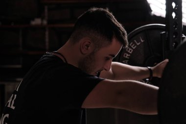 Spor kıyafetleri içindeki dalgın genç erkek sporcunun beden eğitimi sırasında ağır halteri kaldırmaya çalışırken gözleri kapalı uzanan portresi.