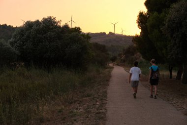 Tanımlanamayan turistlerin sıradan kıyafetleriyle yeşil ağaçların yanında yürürken alacakaranlıkta yeşil tepelerde rüzgar jeneratörleri ile.