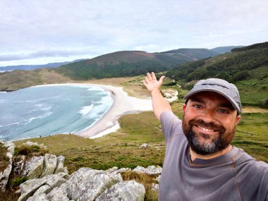 Şapkalı bir adam arka planda Soesto plajı, Laxe, Galiçya ile selfie çekiyor.