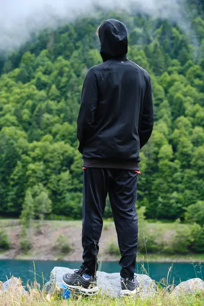 Kapüşonlu, cebinde elleri olan, çimenli tepenin kayalıklarında dikilip yeşil ormanın resimli manzarasına hayran olan isimsiz bir erkek yolcunun arka planı.