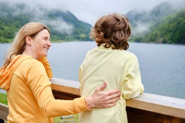 Tahta çitte gülümseyen anne ve çocuğun yan görünüşü. Birbirlerine bakıyorlar ve göl ve dağlara karşı doğanın tadını çıkarıyorlar.