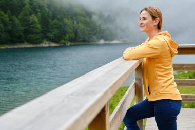 Tırabzana yaslanmış, dağlarda bir göle bakan, tatili sırasında dinlenen bir kadının yan görüntüsü..