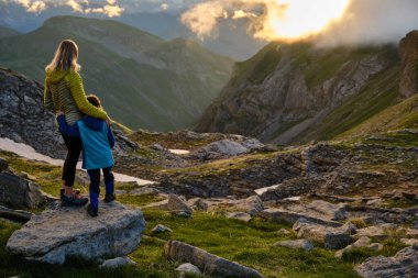 Anne ve oğul gün batımında dağlarda kucaklaşıp yürüyüş sırasında manzarayı izliyorlar..