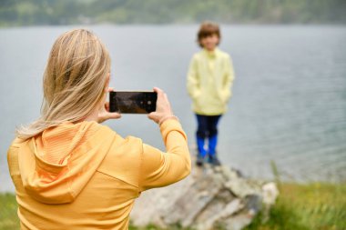 Tanınamayan bir annenin gölün ve dağların önündeki tatillerinde bir çocuğun fotoğrafını çekerken cep telefonu ekranına bakması...