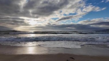 Dalgalar kumlu bir plaja yuvarlanıyor mavi gökyüzünün arka planında dramatik gök gürültülü bulutlar, gün batımı, güzel manzara, 4k ağır çekim