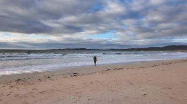 Sörf tahtasıyla siyah dalış kıyafeti giyen bir adam Atlantik Okyanusu 'nda suya giriyor, dikiz manzaralı, yavaş çekim