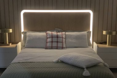 Yatak başlığının üstünde dekoratif ışıklandırma ve yan taraflarında komodinlerle rahat çift kişilik yatak, yatak odası.