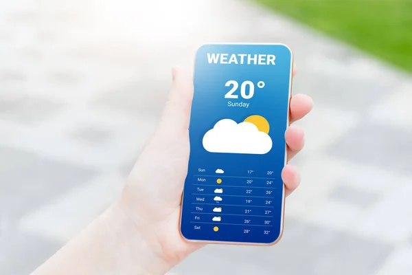 stock image The girl checks the weather forecast in a mobile application. A woman's hand holds a mobile phone and looks at the weather forecast for the day on a park with a trees background