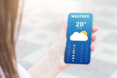 Kız mobil bir uygulamada hava tahminini kontrol ediyor. Bir kadının eli bir cep telefonu tutuyor ve bir parkın arka planındaki hava tahminlerine bakıyor.