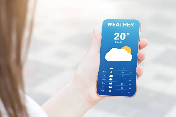 stock image The girl checks the weather forecast in a mobile application. A woman's hand holds a mobile phone and looks at the weather forecast for the day on a park with a trees background