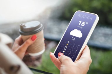 rainy weather. weather forecast for autumn. female hand holds a mobile phone and looks at the weather forecast for the day on a park with a trees background clipart